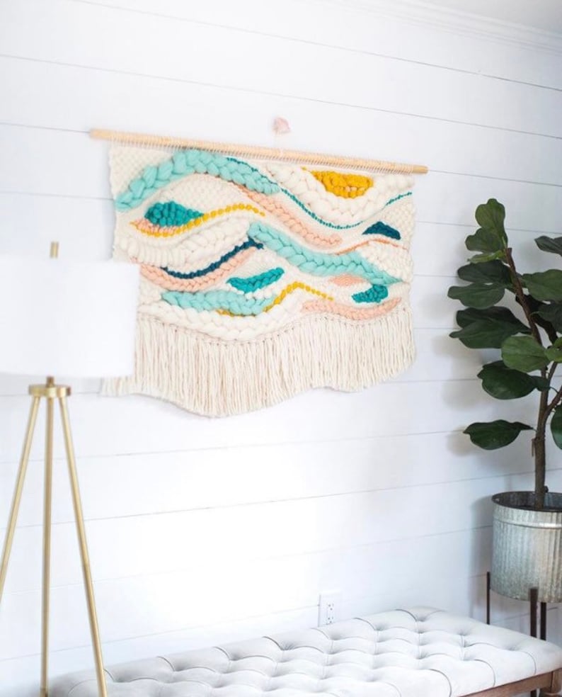 May include: A white woven wall hanging with a wood dowel at the top. The hanging features a wavy pattern in shades of blue, yellow, and pink. The hanging has a fringe at the bottom.