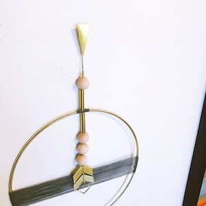 May include: A gold metal wall hanging with a circular frame and a vertical arrow design. The arrow is made of gold metal and has a series of wooden beads hanging from it.
