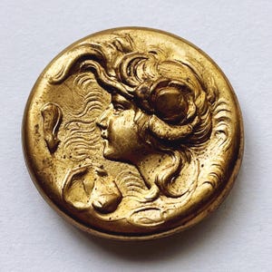 May include: A gold-toned metal button with a raised design of a woman's face with flowing hair. The button has a detailed, ornate design.