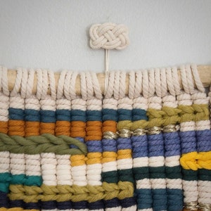Könnte beinhalten: Ein farbenfroher Makramee-Wandbehang mit einem weißen Knoten oben. Der Wandbehang ist aus verschiedenen farbigen Garnen gefertigt, darunter Blau, Grün, Gelb und Orange.