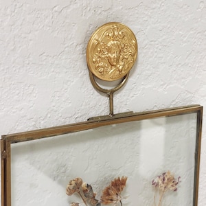 May include: A rectangular glass frame with a brass metal border, featuring a decorative gold-toned circular medallion at the top with a relief of a face surrounded by floral motifs. The frame contains pressed dried flowers in muted brown and purple tones.