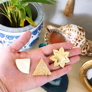 Könnte beinhalten: Drei goldfarbene Metallobjekte: ein Stern, ein Dreieck und ein Halbkreis. Die Objekte liegen auf einer Hand, im Hintergrund sind eine Muschel und eine Topfpflanze zu sehen.