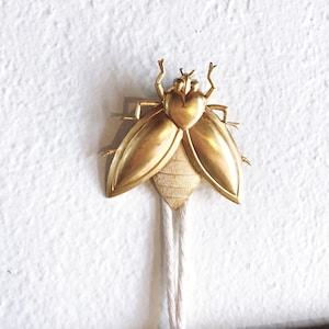 May include: A gold-colored insect-shaped pendant with a white cord. The insect has a heart-shaped body and wings.