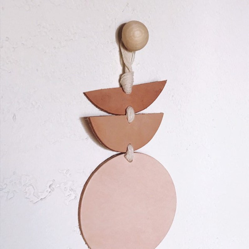 Op de afbeelding: Een wanddecoratie met een houten kraal en drie leren vormen in bruin- en roze tinten. De vormen omvatten twee halve cirkels en een volledige cirkel, verbonden door een natuurlijk koord.