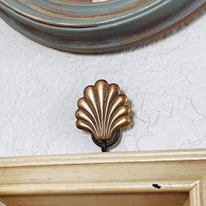 May include: A gold-colored seashell-shaped hook is mounted on a white wall. The hook is attached to a dark metal ring. A light-colored wooden frame is below the hook, and a blue and gold-trimmed oval frame is above.
