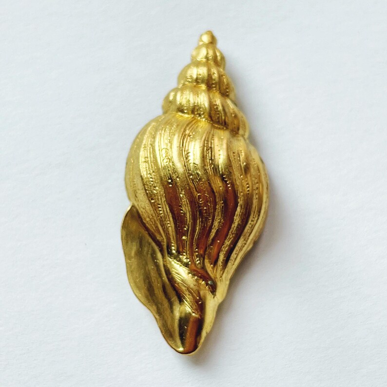 May include: A gold-colored seashell pendant with a detailed, textured surface. The shell is shaped like a spiral and has a pointed tip.