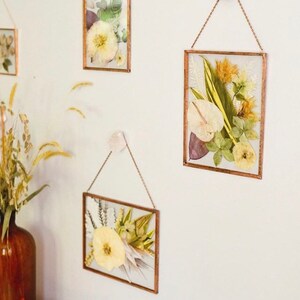 May include: Four framed pressed flower arrangements hanging on a white wall. The frames are made of gold metal and have a simple design. The flowers are a variety of colors, including white, yellow, green, and brown.