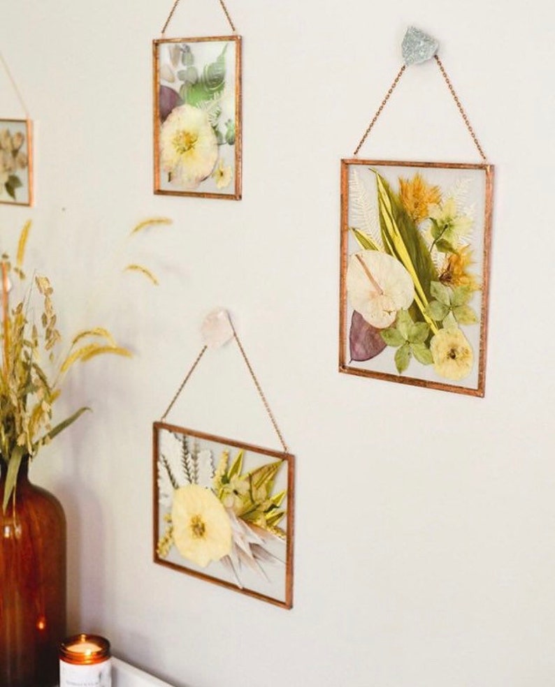 May include: Four framed pressed flower arrangements hanging on a white wall. The frames are made of gold metal and have a simple design. The flowers are a variety of colors, including white, yellow, green, and brown.