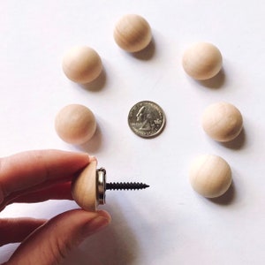 Op de afbeelding: Een verzameling onafgewerkte houten kogelknoppen, met een munt voor schaalvergelijking. E&eacute;n knop is bevestigd aan een schroef, vastgehouden door een hand. De knoppen zijn lichtbruin en gerangschikt op een witte ondergrond.