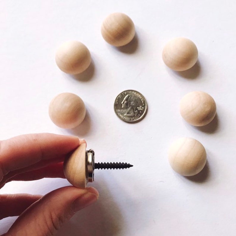 Op de afbeelding: Een verzameling onafgewerkte houten kogelknoppen, met een munt voor schaalvergelijking. E&eacute;n knop is bevestigd aan een schroef, vastgehouden door een hand. De knoppen zijn lichtbruin en gerangschikt op een witte ondergrond.