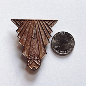 Peut inclure: Broche de style Art Déco de couleur bronze, avec un motif en forme d'éventail. La broche présente des motifs géométriques complexes et est présentée à côté d'une pièce de monnaie pour comparaison de taille. La broche est probablement en métal.