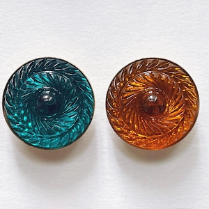 Könnte beinhalten: Zwei runde, dekorative Knöpfe. Einer ist türkis, der andere bernsteinfarben. Beide Knöpfe haben ein Spiraldesign mit einem zentralen, dunkel gefärbten, kreisförmigen Akzent. Der Hintergrund ist weiß.