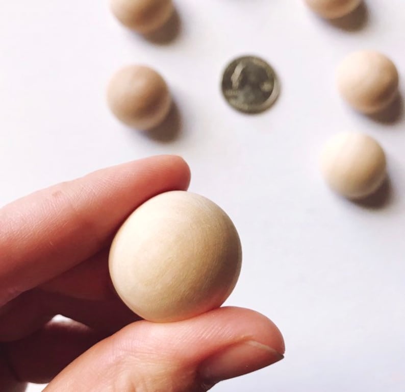 Op de afbeelding: Een close-up van verschillende kleine, lichtgekleurde houten bollen, waarvan er &eacute;&eacute;n tussen de vingers wordt gehouden. Er is ook een munt zichtbaar. De bollen zijn gelijkmatig verdeeld over een wit oppervlak. De houten bollen hebben een diameter van ongeveer 2,5 cm.