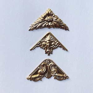 May include: Three gold-toned metal corner brackets with ornate designs. The top bracket features a floral design, the middle bracket features a face, and the bottom bracket features a swirling design.