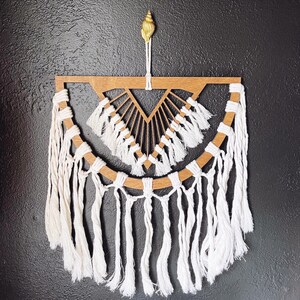 May include: A white macrame wall hanging with a wooden geometric design in the center. The macrame has a fringe border and is hanging from a gold seashell.