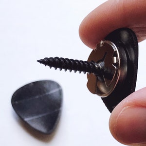 Puede incluir: Una púa de guitarra negra con un tornillo unido a un botón de correa de guitarra negro y plateado.