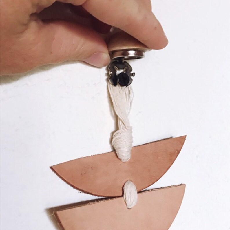 Op de afbeelding: Handgemaakte leren oorbel met een lichtbruine kleur. De oorbel heeft twee halvemaanvormige leren stukken die met een witte draad zijn verbonden. Bevestigd aan een houten en metalen sluiting.
