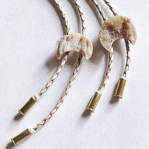 Op de afbeelding: Drie witte leren bolo-strikken met bruine accenten en kristallen maanhangers. De bolo-strikken hebben messing kogelpunten.