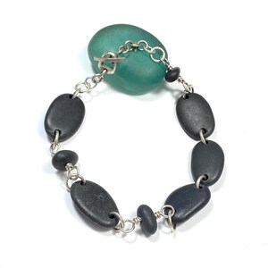 Peut inclure: Un bracelet fait à la main avec des pierres ovales gris foncé lisses reliées par une chaîne argentée. Le bracelet est relié à une pierre de couleur turquoise avec un fermoir à bascule argenté. Le bijou est présenté sur fond blanc.