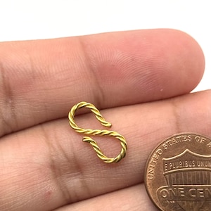 May include: A small, gold-colored S-shaped jewelry clasp with a twisted, rope-like texture. The clasp is positioned next to a United States one-cent coin for scale, highlighting its compact size and detailed metallic finish.