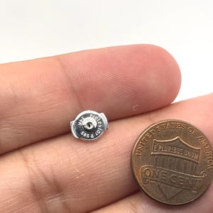 May include: A silver-colored earring back with a circular shape and a central hole. The metal surface features the engraved text 'Protektor', '14K', and '585'. It is positioned next to a United States one-cent copper coin for scale reference.