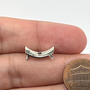 May include: A small, curved silver-toned metal component featuring two downward-pointing prongs and a tiny, centered rectangular red stone. The object is positioned next to a United States one-cent coin for scale, showing its compact size.