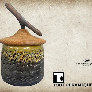 Peut inclure: Un pot en céramique artisanal avec un couvercle et une poignée en bois. Le pot a une surface texturée avec un dégradé de couleurs, passant du gris foncé au vert olive. Le couvercle est en bois brun clair. Le texte "100% Fait main au Québec" est visible.