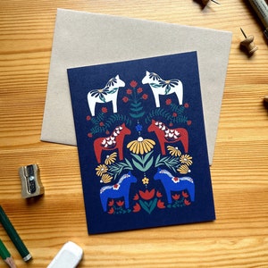 May include: A navy blue greeting card featuring a folk art design of horses and floral elements in red, white, yellow, and blue. The card is paired with a tan envelope, and is on a wooden surface.