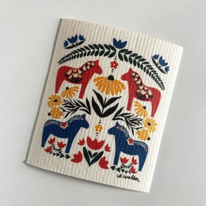 May include: A white dishcloth with a colorful Scandinavian folk art design featuring red and blue stylized horses, yellow flowers, and green leaves. The design is framed by a border of green leaves and blue flowers. The text "c.e. walker" is printed in the bottom right corner.