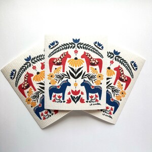 May include: Three white dishcloths with a Scandinavian folk art design featuring red and blue horses, yellow flowers, and green leaves. The dishcloths are stacked on top of each other.
