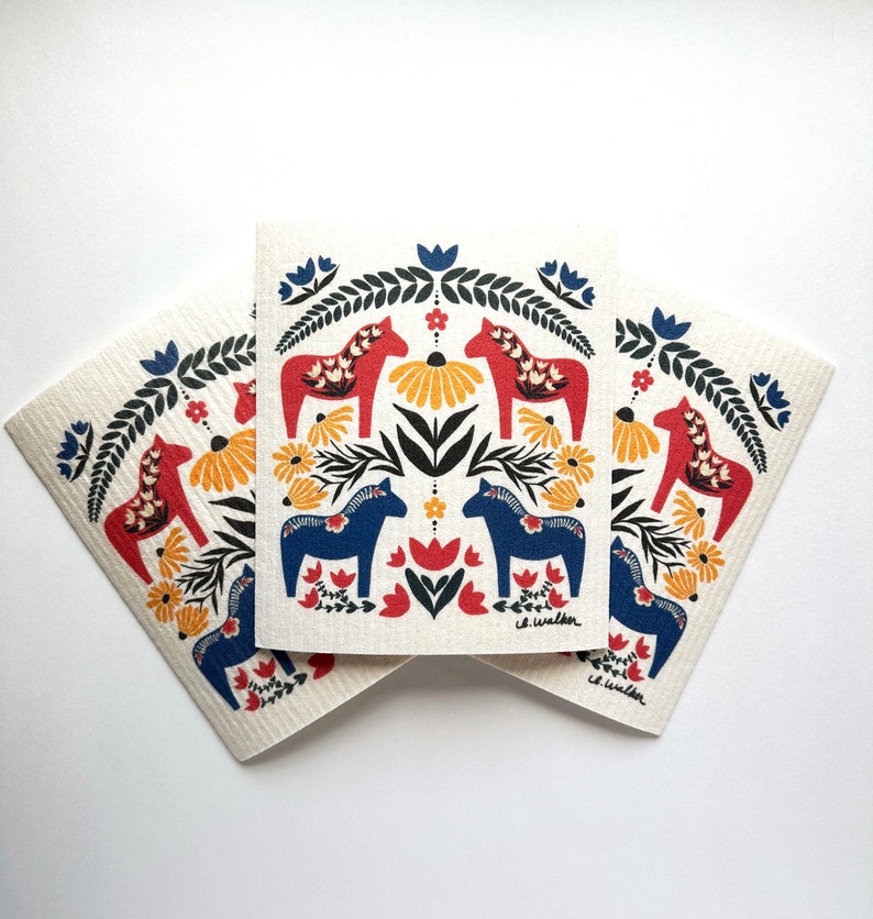 May include: Three white dishcloths with a Scandinavian folk art design featuring red and blue horses, yellow flowers, and green leaves. The dishcloths are stacked on top of each other.