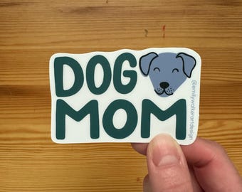 May include: A white sticker with the words "DOG MOM" in dark teal letters. A light blue dog face is on the right side. The sticker is held by a hand.