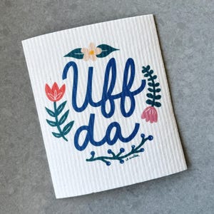 May include: White rectangular cleaning cloth with the blue text "Uff da" surrounded by floral illustrations in red, pink, and green. The cloth has a textured surface and is on a gray background.