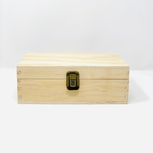 May include: A natural wood box with a brass latch. The box is closed and has a light wood grain.