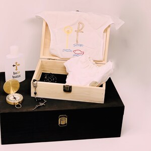May include: A wooden box with a white garment inside, a bottle of holy water, a compass, and a rosary. The garment has an embroidered design of a cross and a fish.