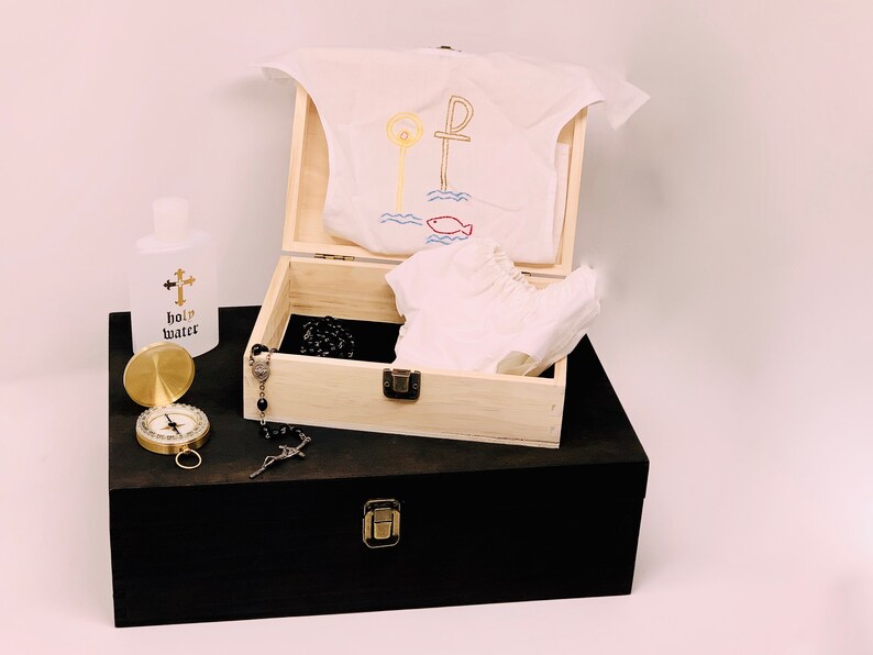 May include: A wooden box with a white garment inside, a bottle of holy water, a compass, and a rosary. The garment has an embroidered design of a cross and a fish.