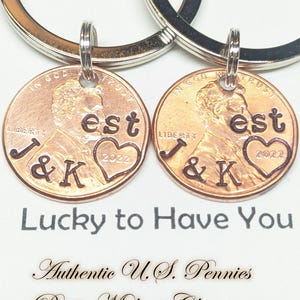 May include: Two copper penny keychains with the text "est", a heart, the initials "J & K", and the year "2022". The keychains are attached to silver key rings. The text "Lucky to Have You" is below the keychains.