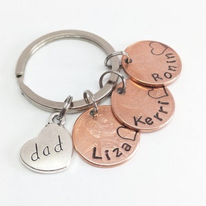May include: A personalized keychain with a silver heart charm that says "dad". The keychain includes four copper pennies with names and hearts, and a silver keyring. The names "Liza", "Kerri", and "Ronin" are visible.
