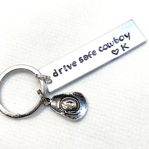 May include: A silver-colored keychain with a rectangular tag that reads "drive safe cowboy" with a heart and the letter "K". A small, detailed cowboy hat charm hangs below the tag.