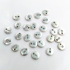 May include: A collection of silver-toned metal alphabet letter charms. The charms are round and have a smooth, shiny finish. Each charm features a single letter of the alphabet in a black font.