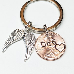 May include: A silver-colored keychain with two angel wing charms and a copper penny. The penny is stamped with the word "Dad", a heart, and the year 2022. The keychain is on a silver-colored ring.