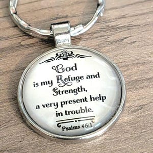 May include: A round silver keychain featuring a glass cabochon with a white background and black text that reads: God is my Refuge and Strength, a very present help in trouble. Psalms 46:1. The metal frame has a polished finish and a split ring.
