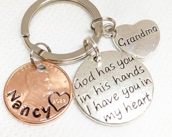 Può includere: Un portachiavi argentato con un ciondolo a forma di cuore che dice "Nonna", un ciondolo argentato con il testo "Dio ti ha nelle sue mani Ti ho nel mio cuore", e una moneta di rame con l'anno 2022 e il nome "Nancy" inciso su di essa.