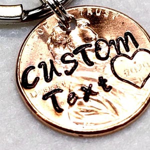 May include: A copper penny keychain with the words "CUSTOM Text" stamped on it in black. A heart with the year "2020" is also stamped on the penny. The keychain has a silver-colored ring.