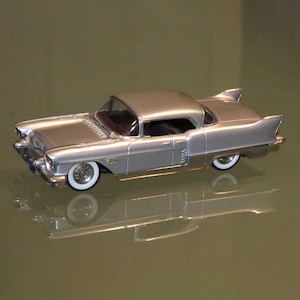 Könnte beinhalten: Silbernes Modellauto mit Weißwandreifen. Das klassische Auto hat ein Hardtop und Chromdetails. Das Auto ist ein Vintage-Modell, wahrscheinlich aus der Mitte des 20. Jahrhunderts, und wird auf einer reflektierenden Oberfläche präsentiert.