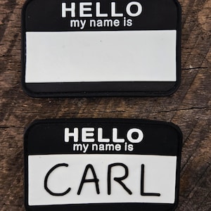 May include: Two rectangular name tag patches with a black border and background. The top portion features the white text HELLO my name is, while the bottom portion is a white rectangular space. The bottom patch has the name CARL written in black.