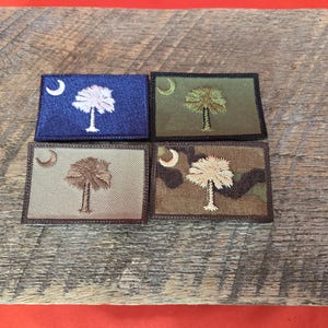 Puede incluir: Cuatro parches bordados con el diseño de la bandera del estado de Carolina del Sur. Cada parche rectangular muestra una luna creciente y una palmera. Los parches son de varios colores, incluyendo azul, verde, beige y camuflaje.