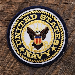 Op de afbeelding: Een ronde geborduurde patch met het embleem van de United States Navy. Het ontwerp bevat een centrale adelaar met gespreide vleugels, een schild en een gouden touwrand op een witte achtergrond met donkerblauwe tekst UNITED STATES NAVY.
