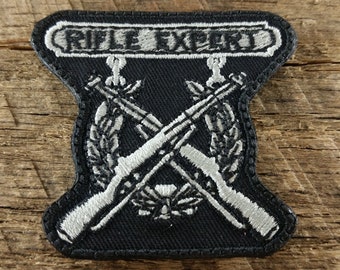 May include: A black embroidered patch with the text "Rifle Expert" in white. Two crossed rifles are below the text, with a wreath around them. The patch has a white stitched border.