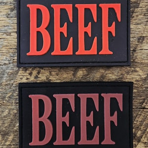 Op de afbeelding: Twee rechthoekige zwarte patches met het woord BEEF in reliëf. De bovenste patch heeft felrode letters, terwijl de onderste patch hetzelfde woord in een donkerdere, gedempte bordeauxrode tint op een zwarte achtergrond weergeeft.